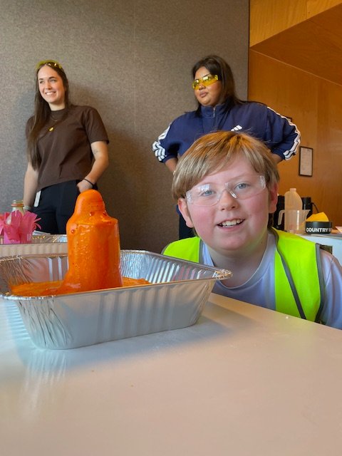 A child smiles beside a foaming volcano experiment with staff nearby